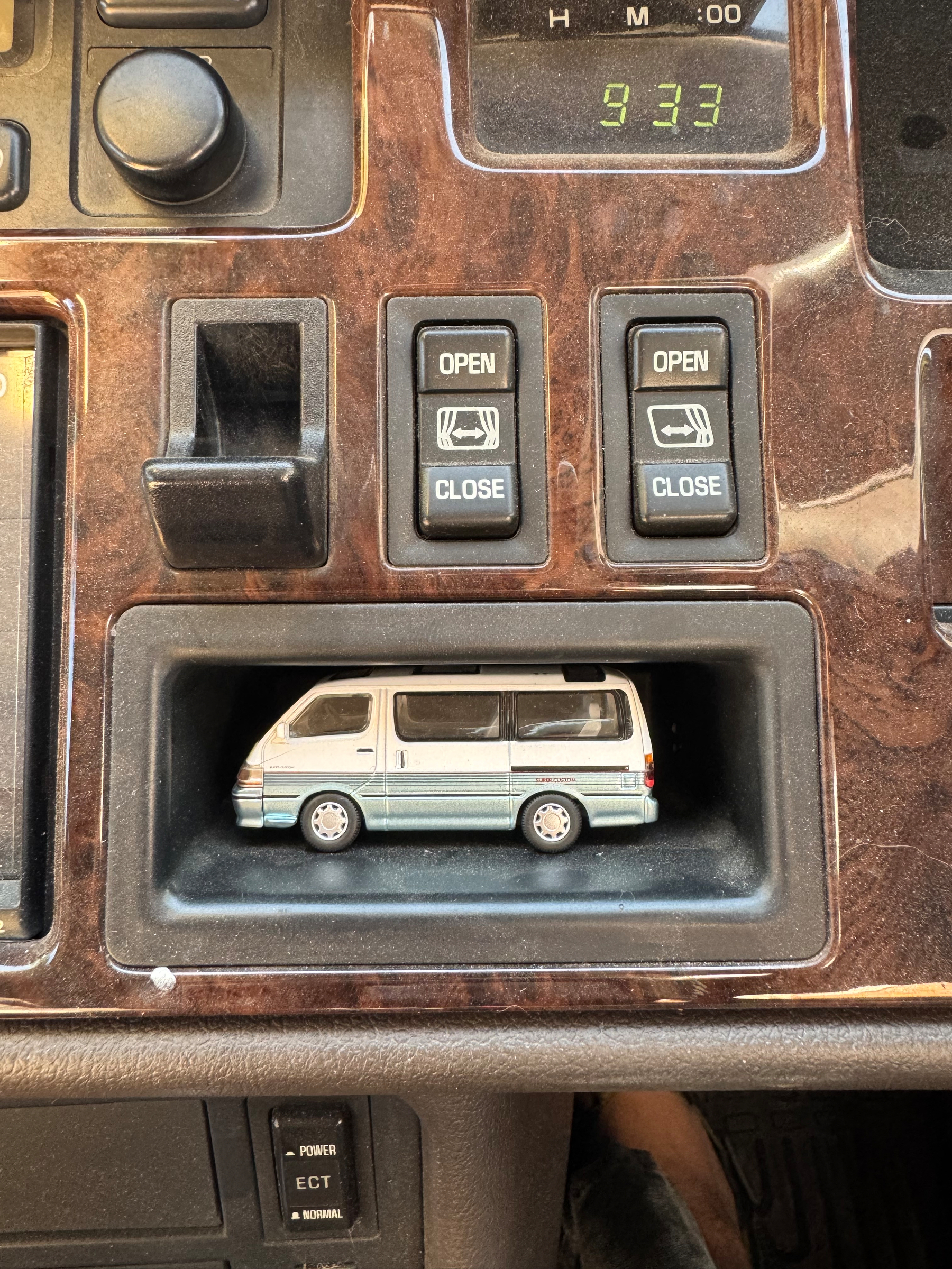 A toy van is placed inside a compartment on a car dashboard that includes controls for opening and closing windows or sunroofs, as well as a digital clock displaying 9:33.