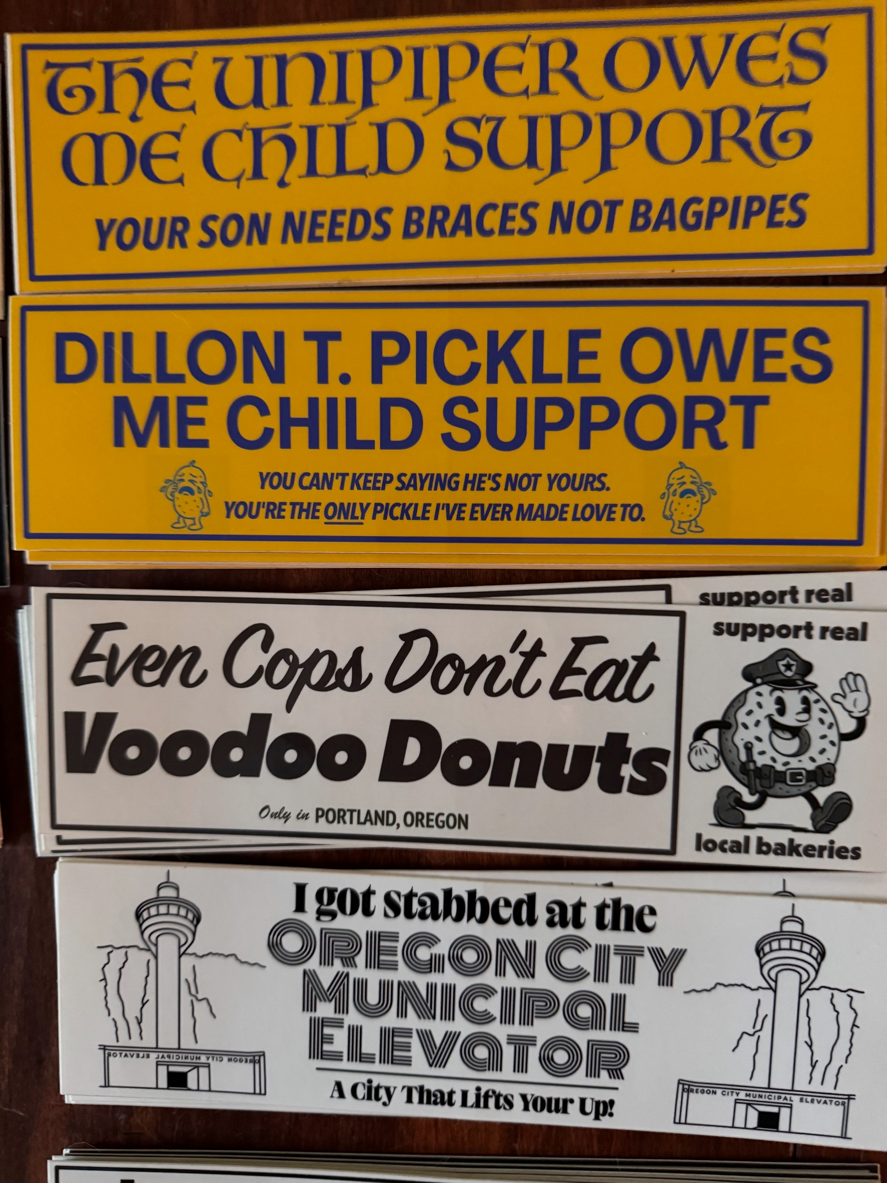Four bumper stickers are displayed, containing humorous and satirical messages about child support, police eating Voodoo Donuts, and an incident at the Oregon City Municipal Elevator.