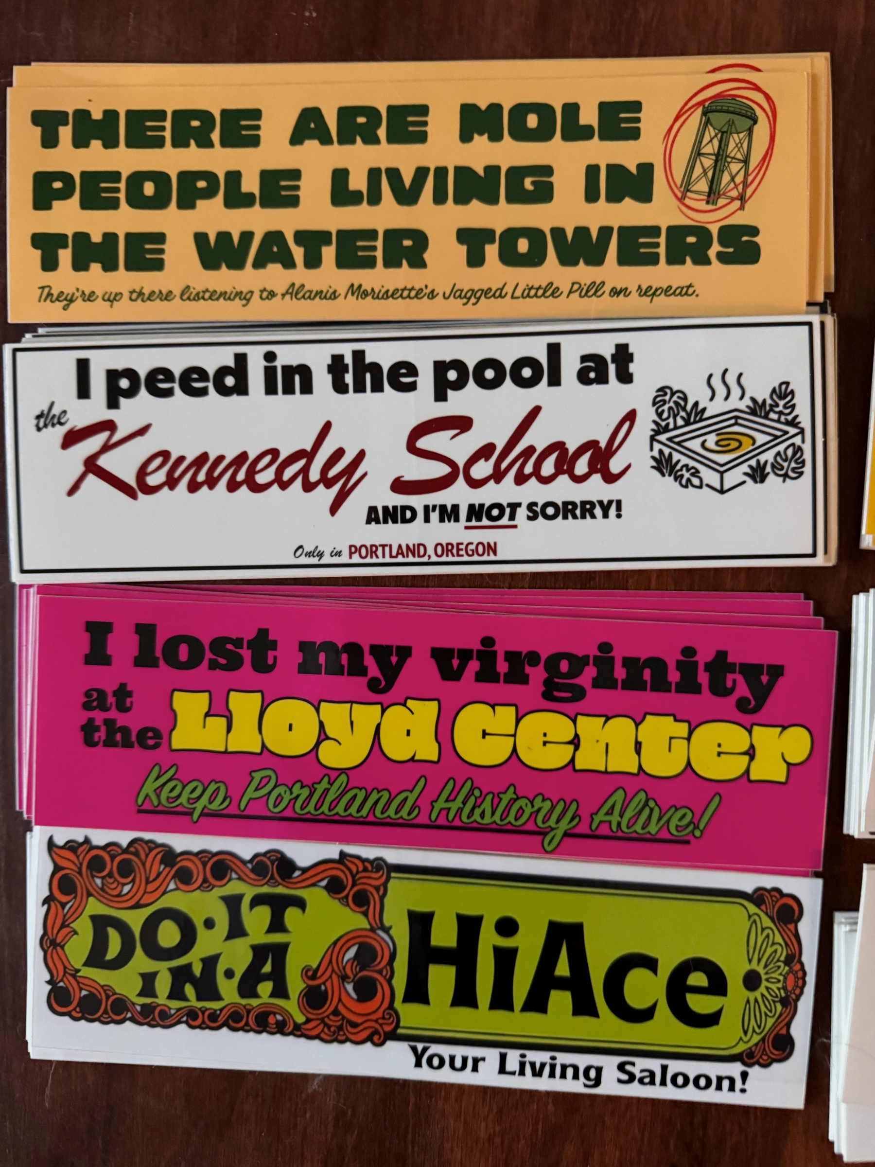 Four colorful bumper stickers with humorous and nostalgic phrases related to Portland, Oregon, are arranged on a wooden surface.