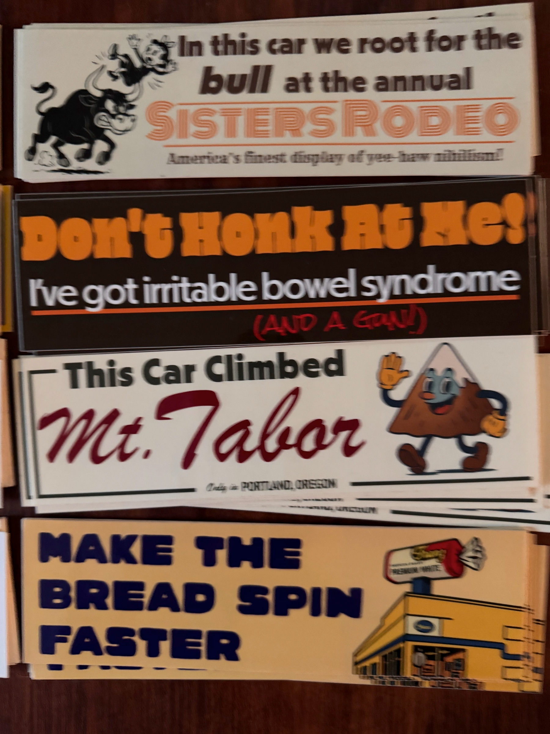 Four humorous and expressive bumper stickers are displayed, featuring themes from rodeo, irritable bowel syndrome, a mountain climb, and a playful slogan about bread spinning.