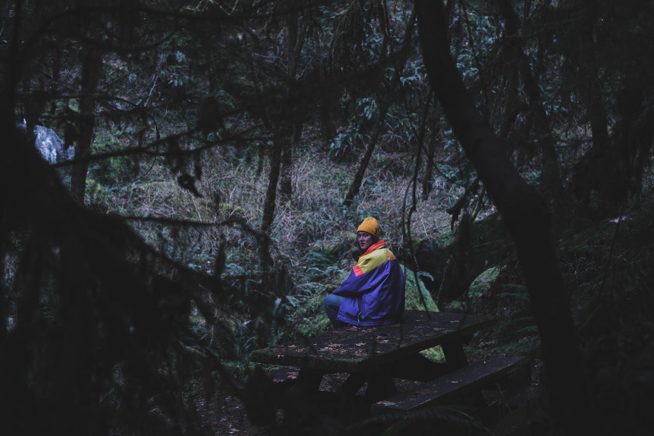 A person wearing a colorful jacket and yellow beanie sits at a picnic table in a dark, forested area. (Oregon Coast, 2022)