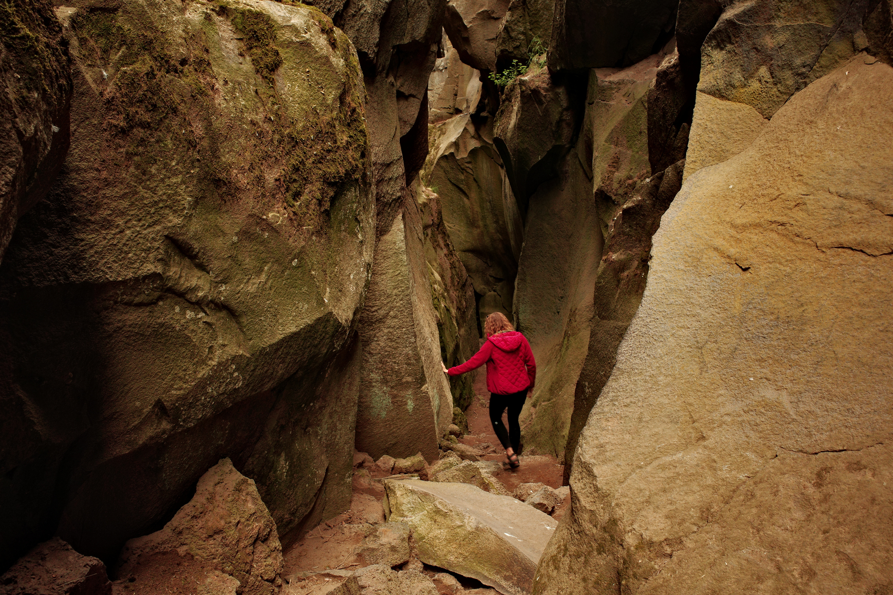 A woman in a red jacket faces away from the camera, walking down a valley created by large jagged rocks.
