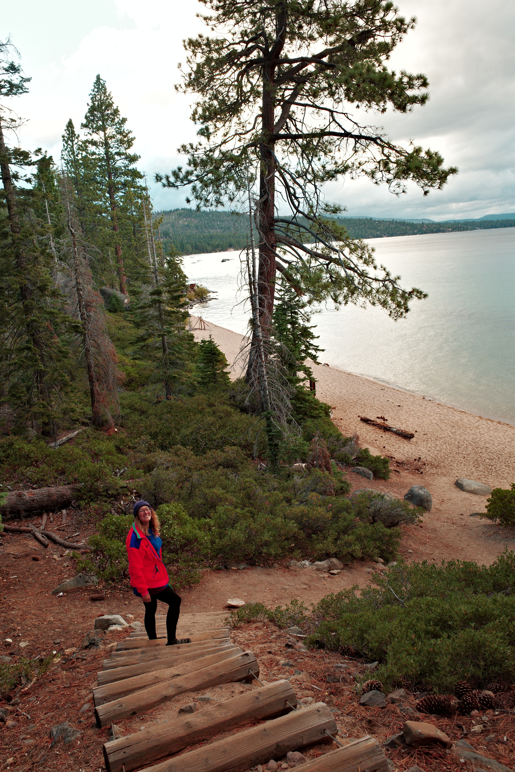 A person wearing a red jacket and a beanie is standing on wooden steps surrounded by pine trees overlooking a sandy beach and calm lake under a cloudy sky.