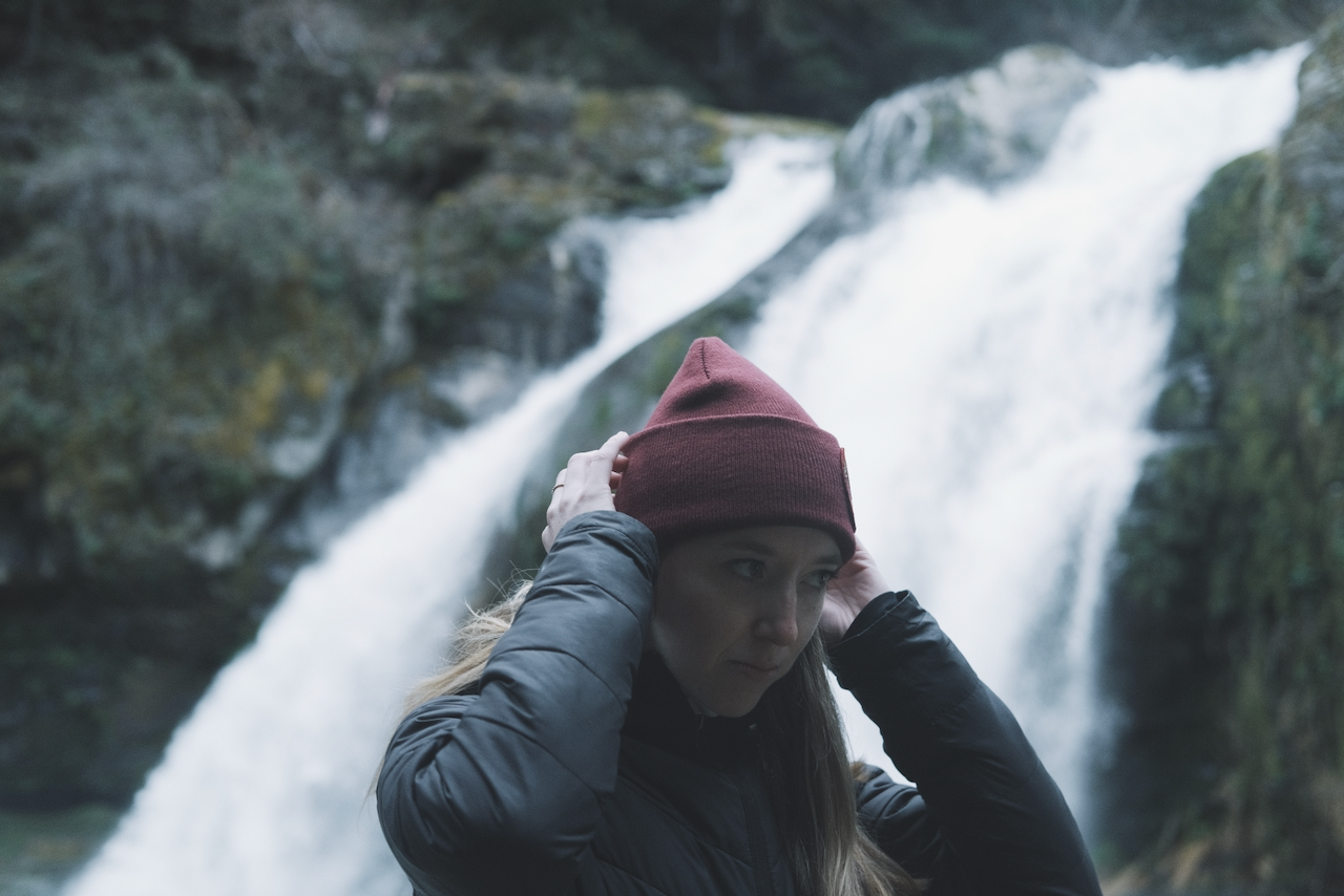 A person wearing a red beanie and a dark jacket stands in front of a waterfall. (Oregon Coast, 2022)