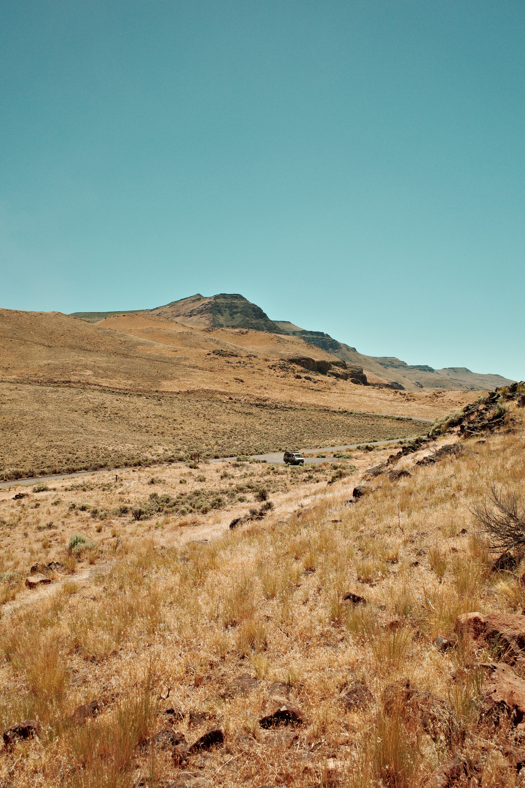 A solitary vehicle is driving along a winding road through a dry, grassy, mountainous landscape under a clear blue sky.