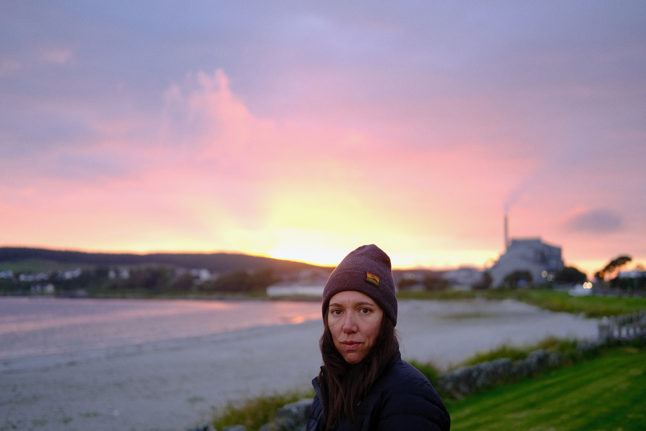 A person wearing a beanie stands near a serene beach at sunset with buildings and smokestacks visible in the background. (Scotland, 2022)