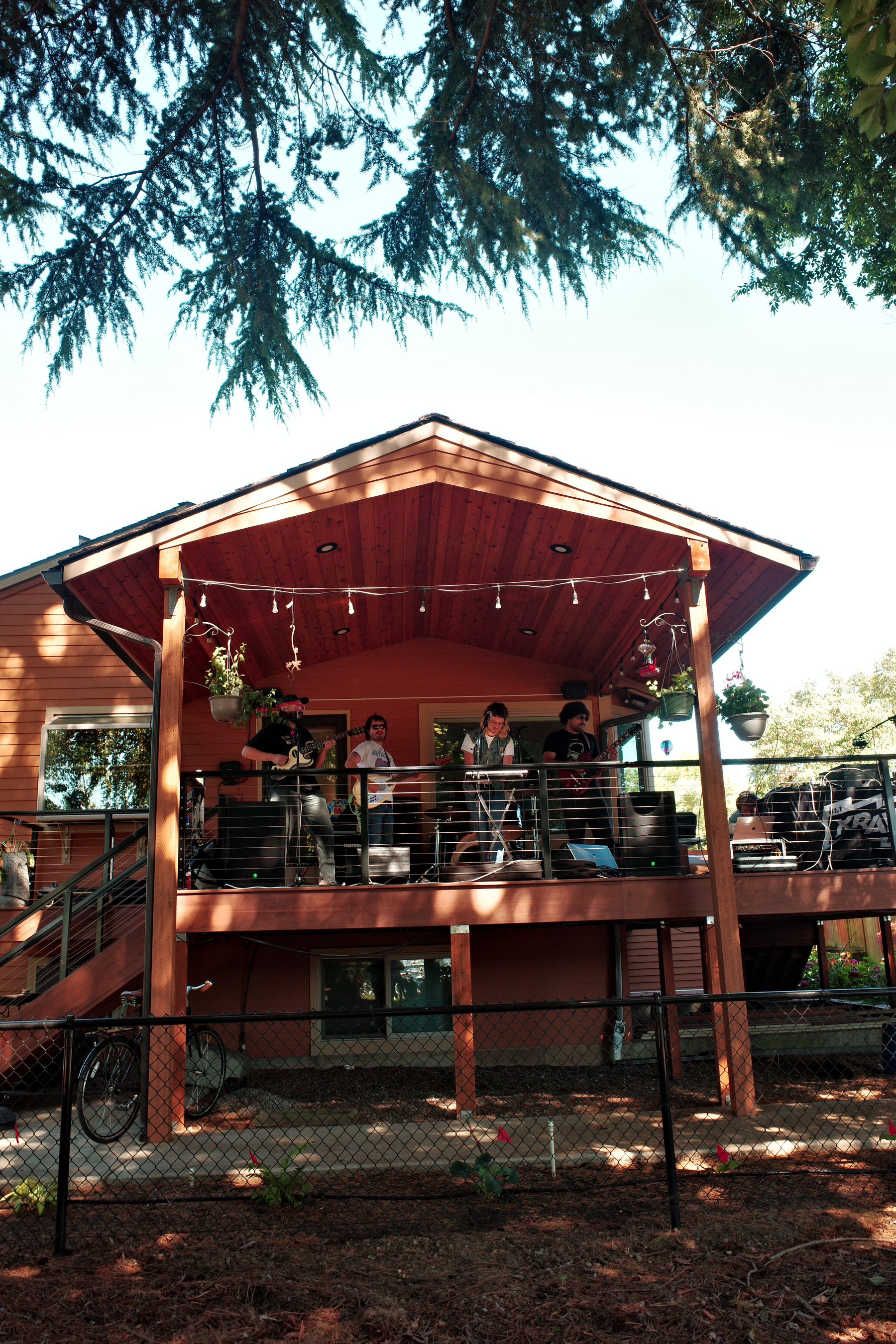 A group of people are playing musical instruments on a wooden porch attached to a house with string lights and hanging plants.