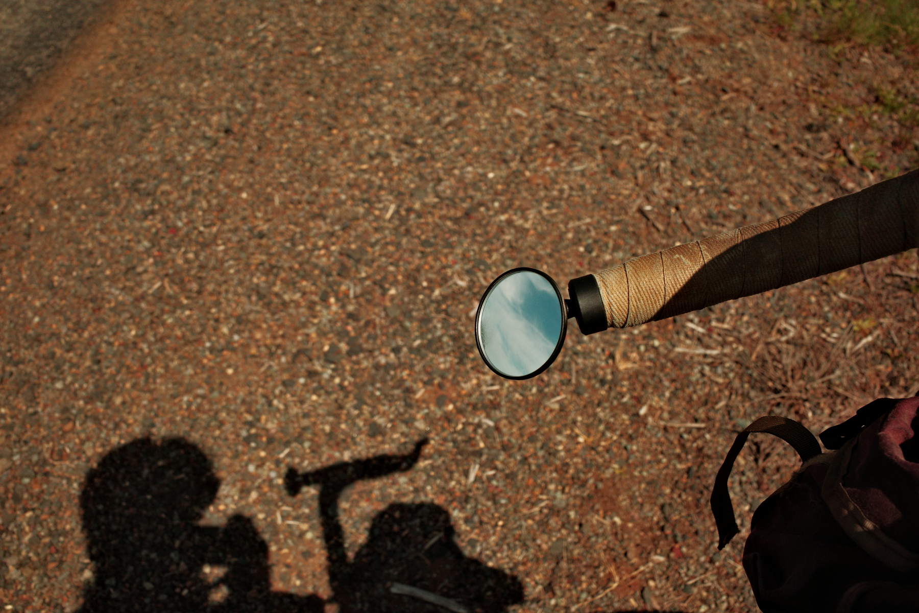A bicycle handlebar with a round mirror reflects the sky, while its shadow is cast on a gravel path.