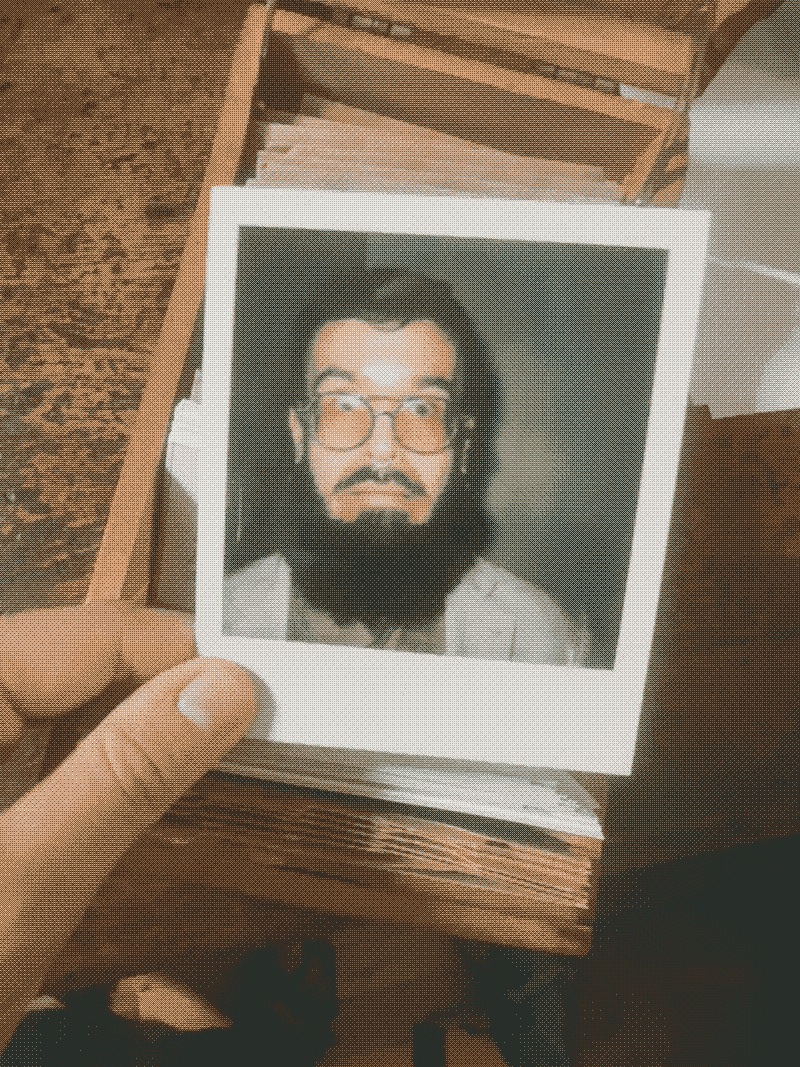 A hand holding a Polaroid photo of a man with glasses and a beard, a wooden box of more photos behind it.