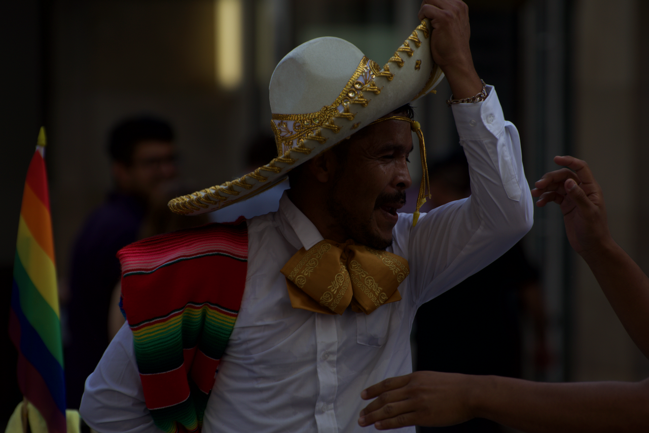 A man wearing a traditional Mexican hat and colorful attire is dancing near a multicolored flag. (New Haven, CT 2018)