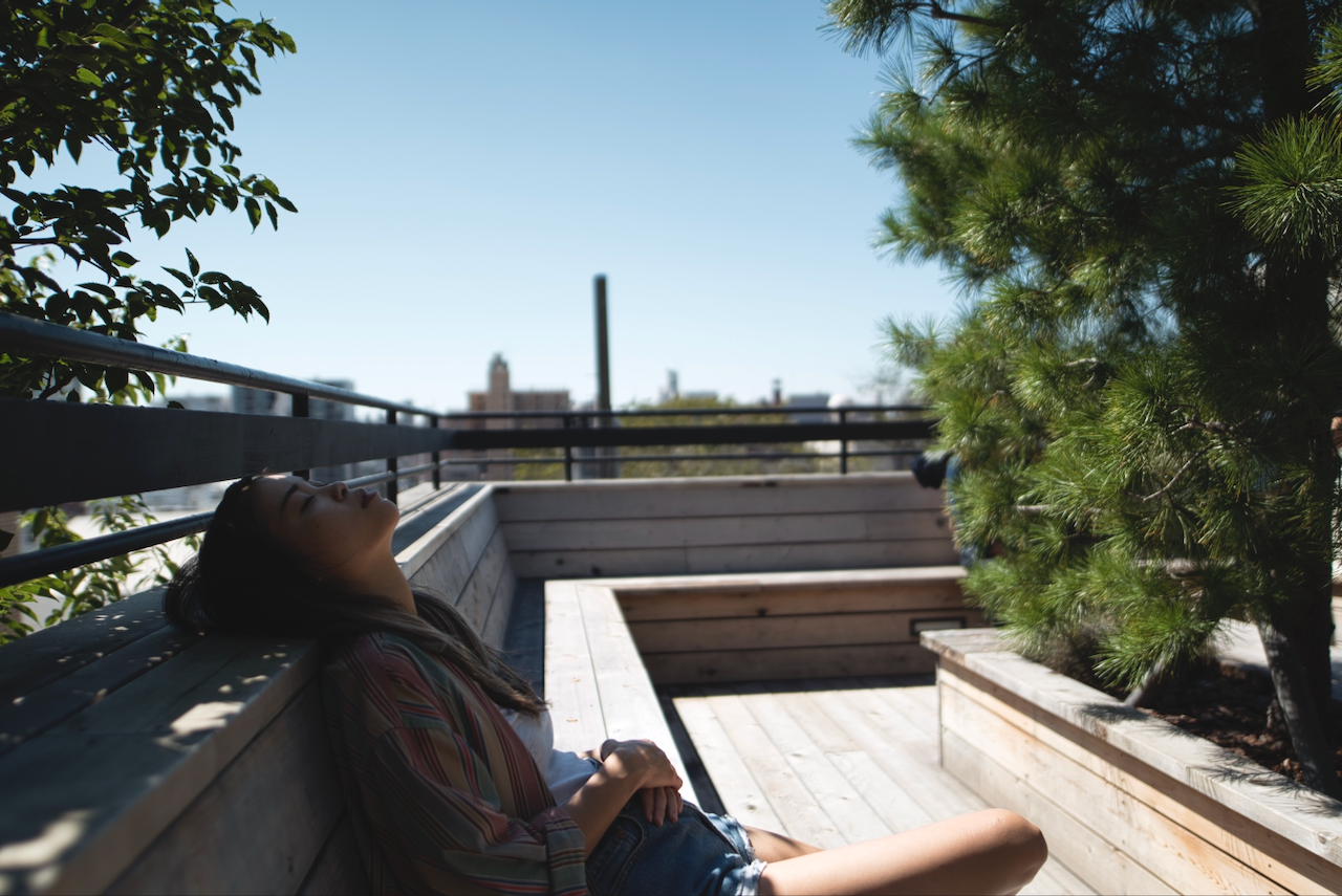 A person is relaxing on a wooden bench in a sunny, outdoor setting surrounded by greenery. (Tokyo, 2018)
