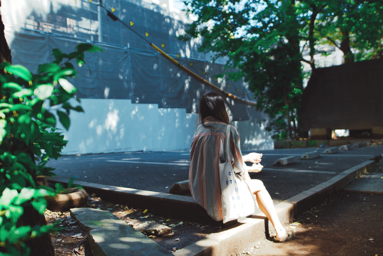 A person is sitting on a curb in a sun-dappled area with trees and construction scaffolding nearby. (Tokyo, 2018)