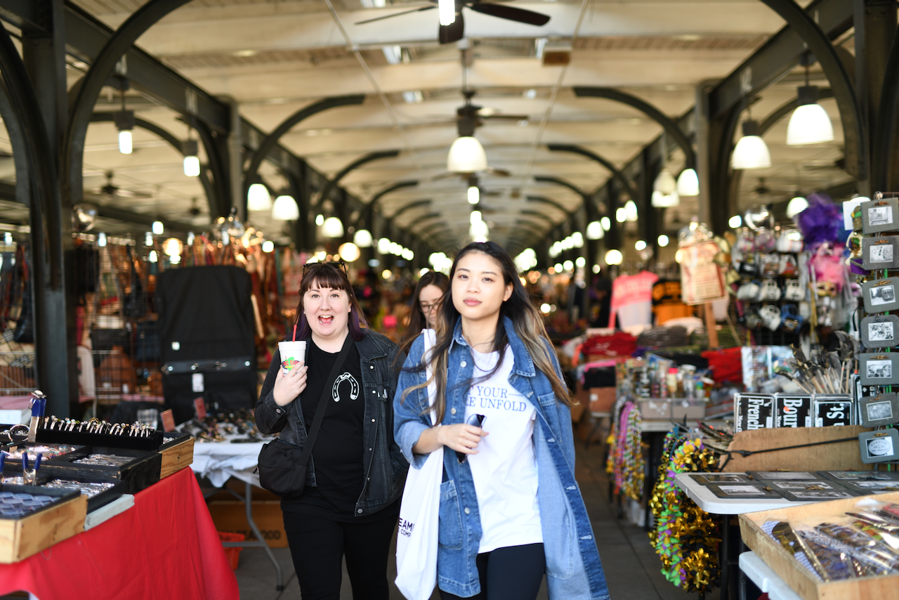 Two women are walking through a vibrant market with various stalls displaying goods. (New Orleans, 2018)