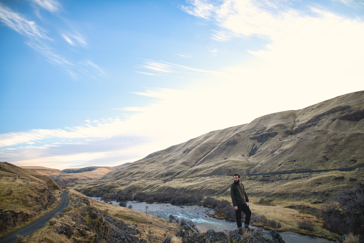 A person stands near a river amidst rolling hills under a bright blue sky with scattered clouds. (Oregon, 2019)