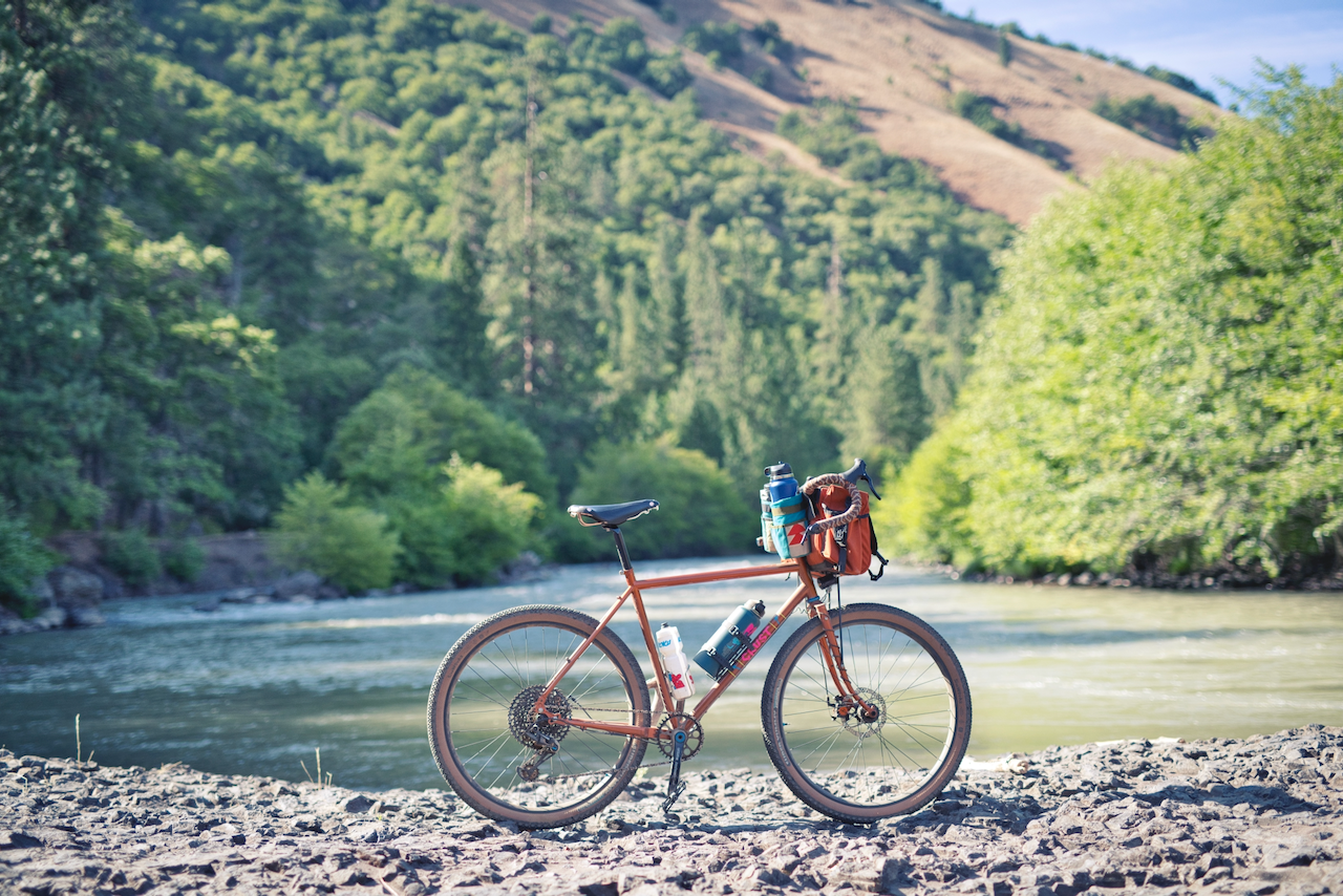 An orange bicycle propped up on the rocky beach of a river. (Klickitat County, WA. 2020.)
