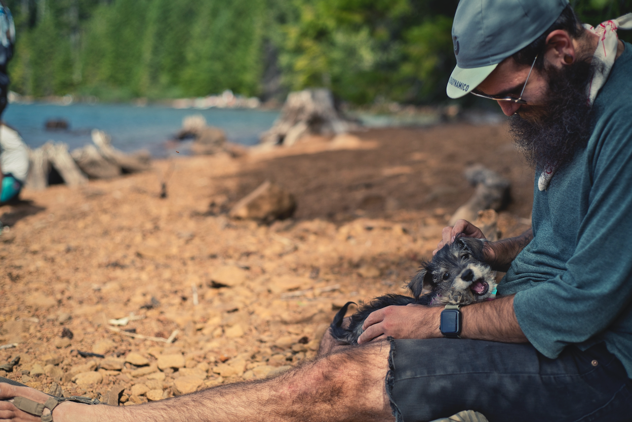 A man with a beard and a cap is sitting on a rocky lakeshore, petting a small dog on his lap. (Mt. Hood, 2020)