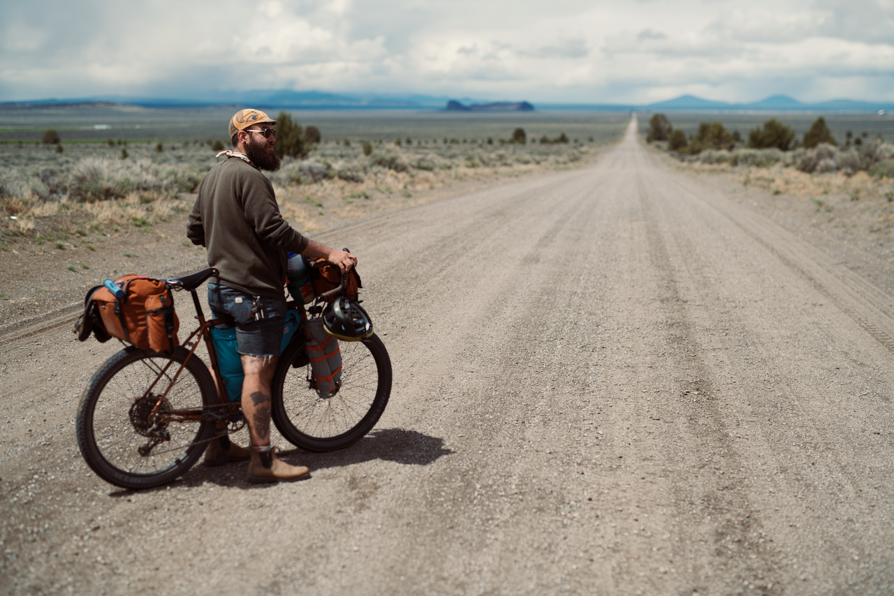 A person with a bicycle stands on a long, gravel road surrounded by an expansive rural landscape. (Ft. Rock, 2020)