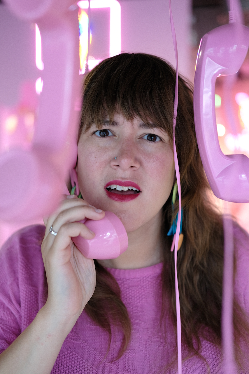 A person with a perplexed expression holds a pink telephone receiver to their ear amidst several hanging pink phones. (Stockholm, 2022)