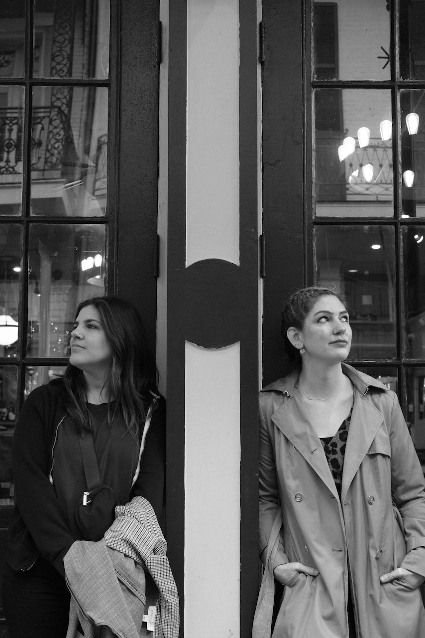 Two women stand back-to-back against a building's doorway, gazing in opposite directions in a black and white photo. (New Orleans, 2023)