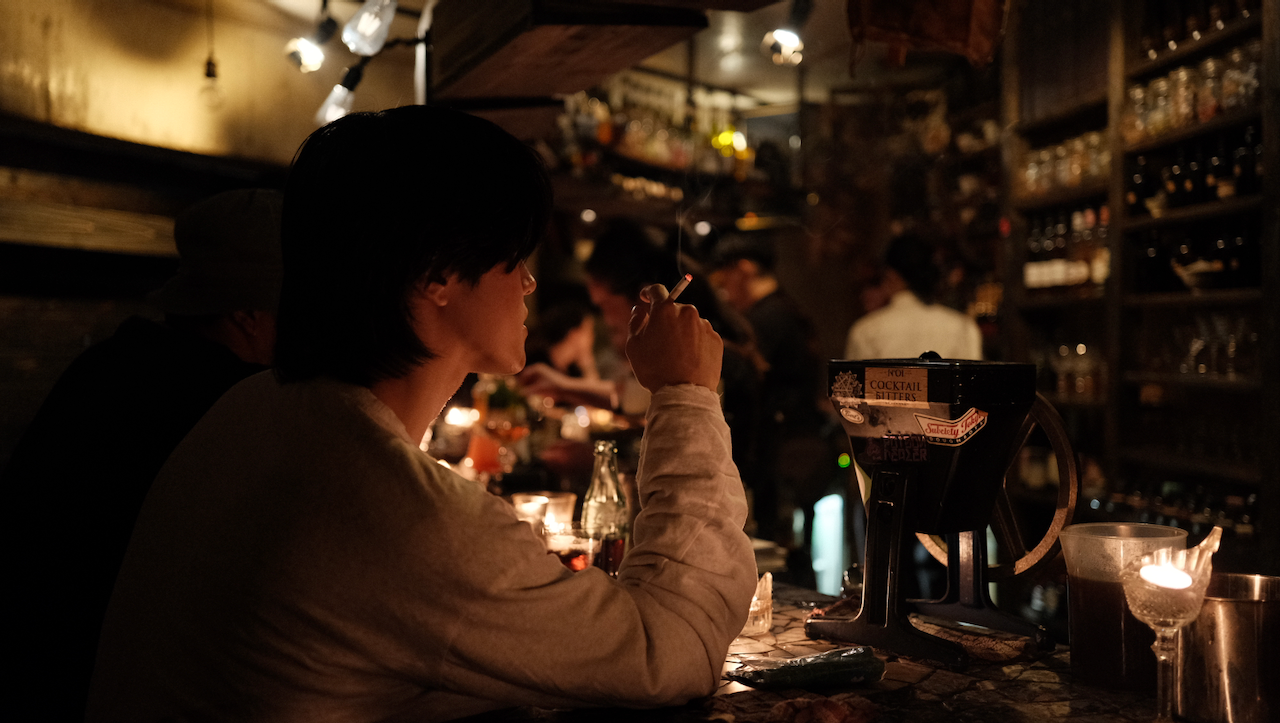 A person is sitting at a dimly lit bar holding a cigarette, surrounded by bottles and warm lighting. (Osaka, 2024)