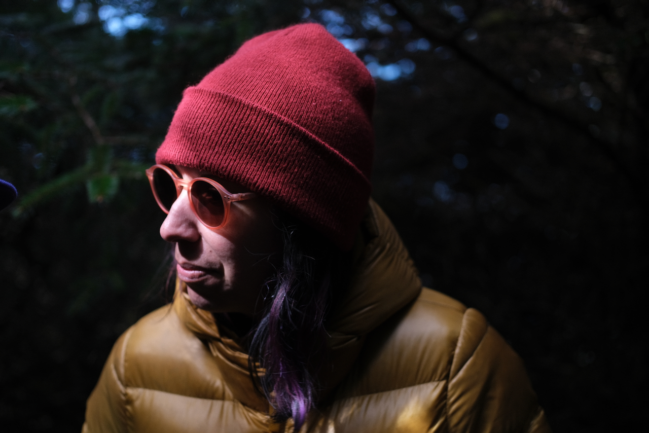 A person wearing a red beanie, round sunglasses, and a yellow puffer jacket is outdoors against a dark, wooded background. (Oregon Coast, 2022)