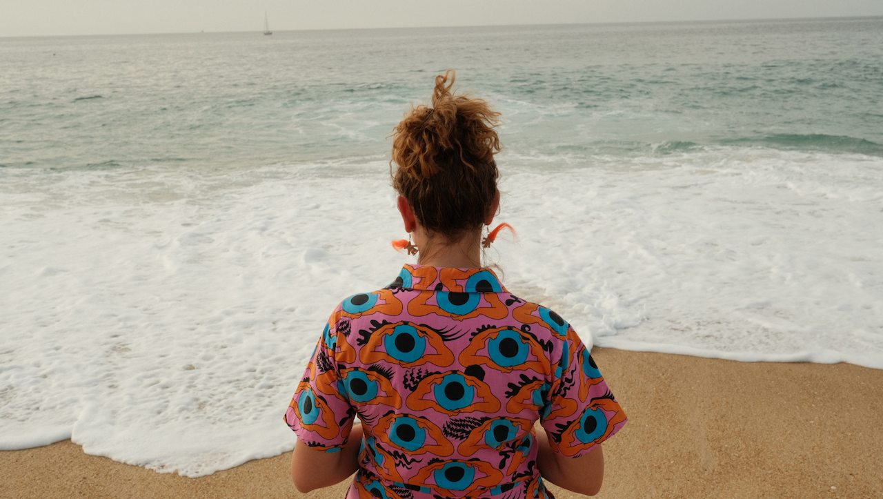 A person with curly hair, wearing a colorful shirt with bold patterns, stands on a beach facing the ocean. (Portugal, 2024)