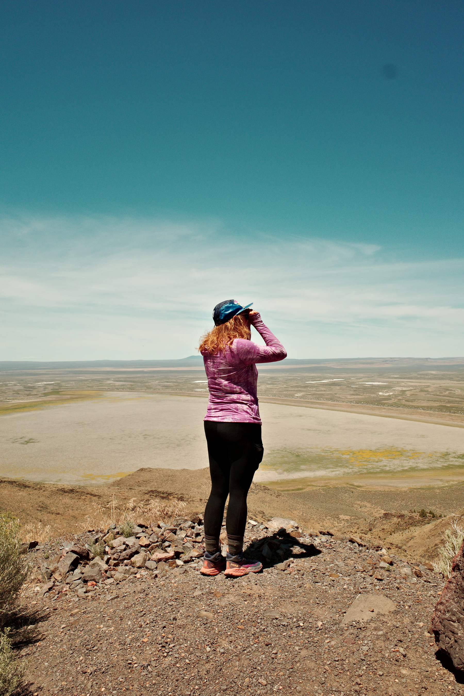 A person wearing a purple long-sleeve shirt, black leggings, and a blue cap is standing on rocky terrain while looking out over a vast, open landscape under a clear sky.