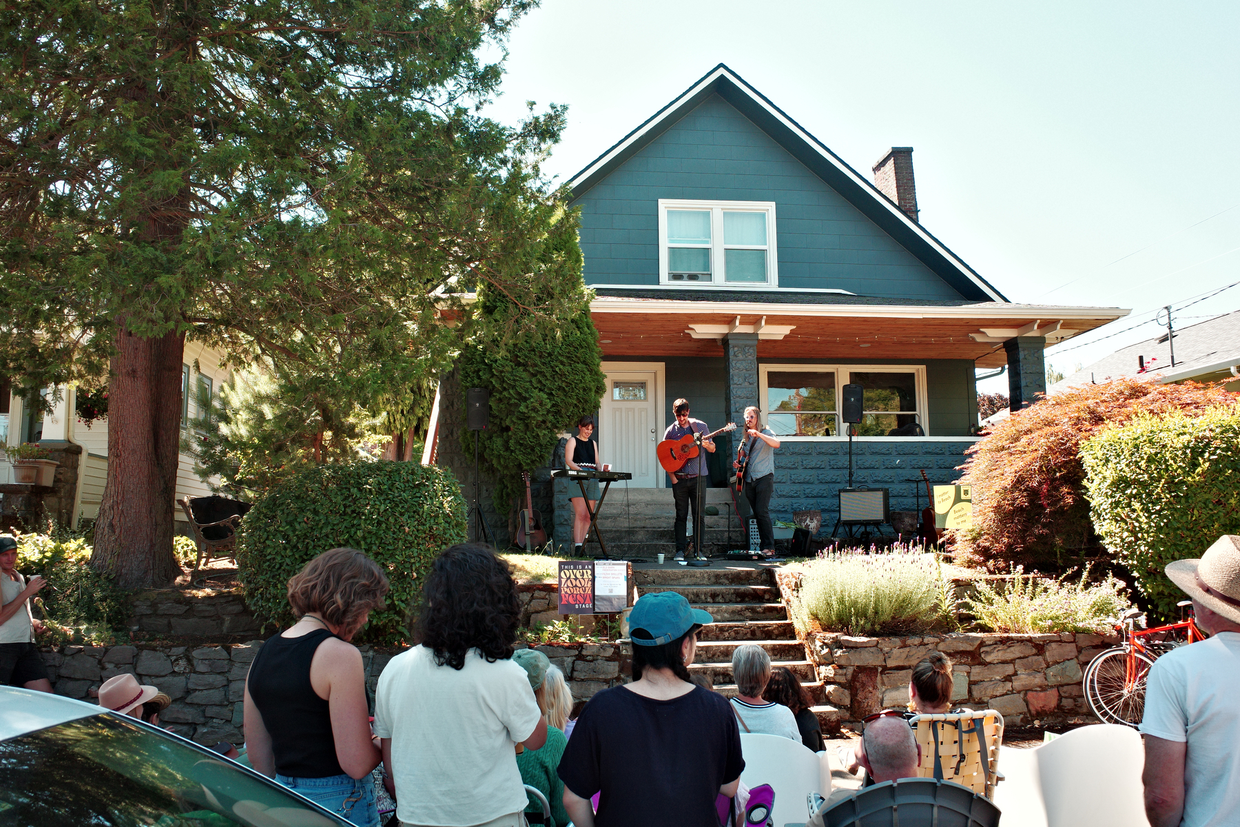 A small group of musicians is performing on the porch of a blue house while an audience watches from the yard and street.