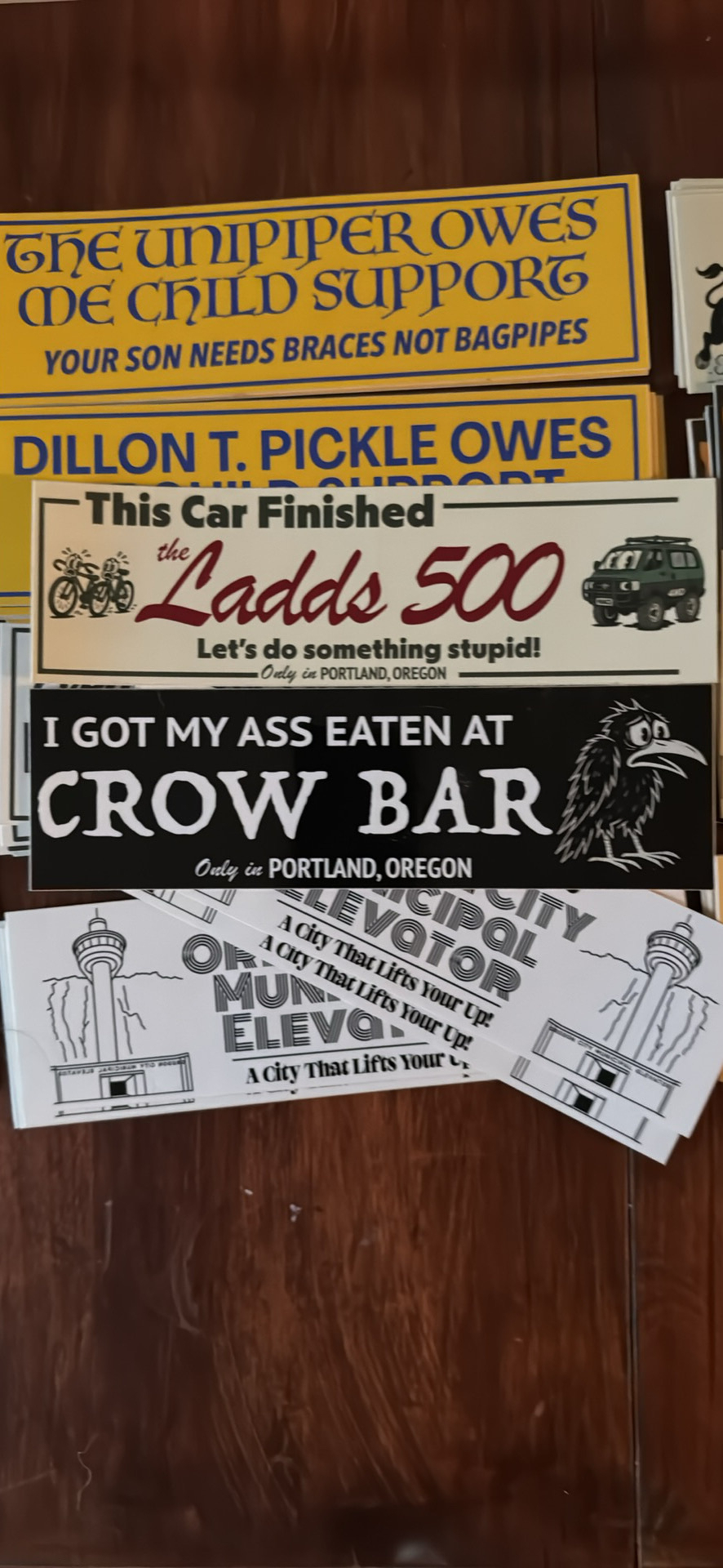 A collection of humorous bumper stickers is displayed on a wooden surface, featuring various quirky phrases about child support, a car race, a bar experience, and Portland, Oregon.