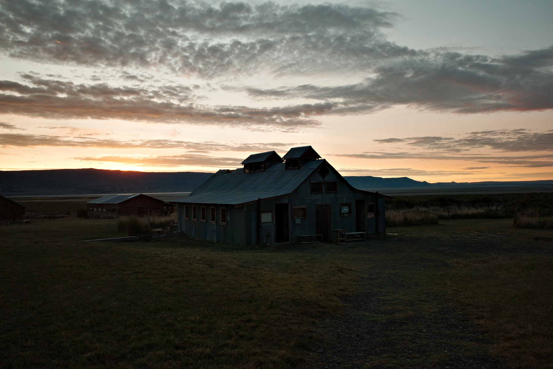 A rustic building with two dormer windows stands in an open field against a backdrop of a sunset sky and distant mountains.
