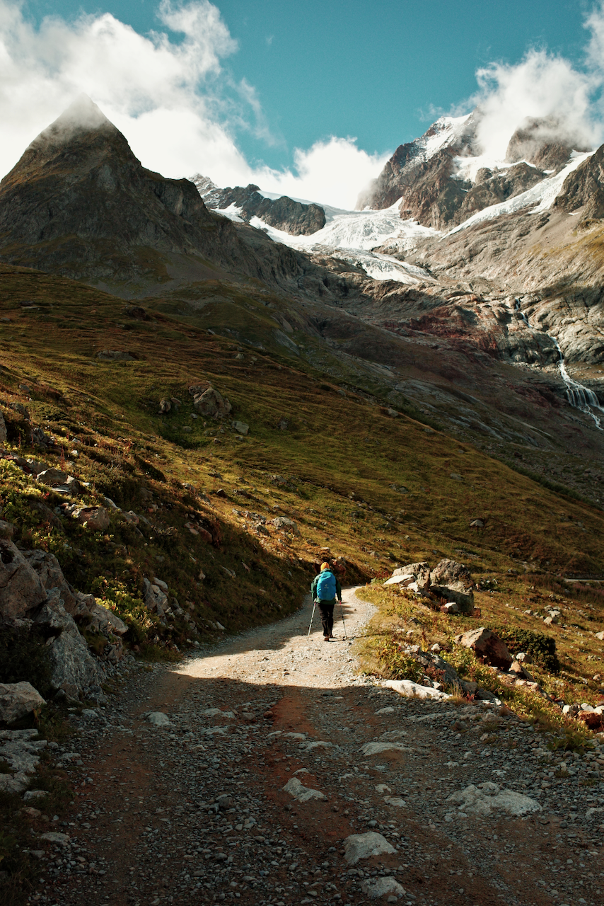 A lone hiker walks along a rugged mountain path under a bright sky, with snow-capped peaks in the background. (Italy, 2025)