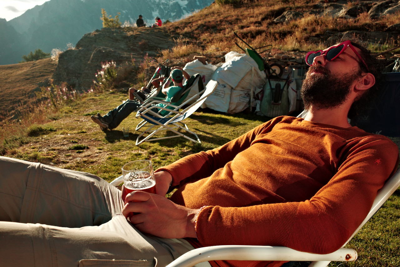Two people relax on outdoor lounge chairs in a sunny, mountainous landscape, one of them wearing heart-shaped sunglasses and holding a drink. (Italy, 2025)