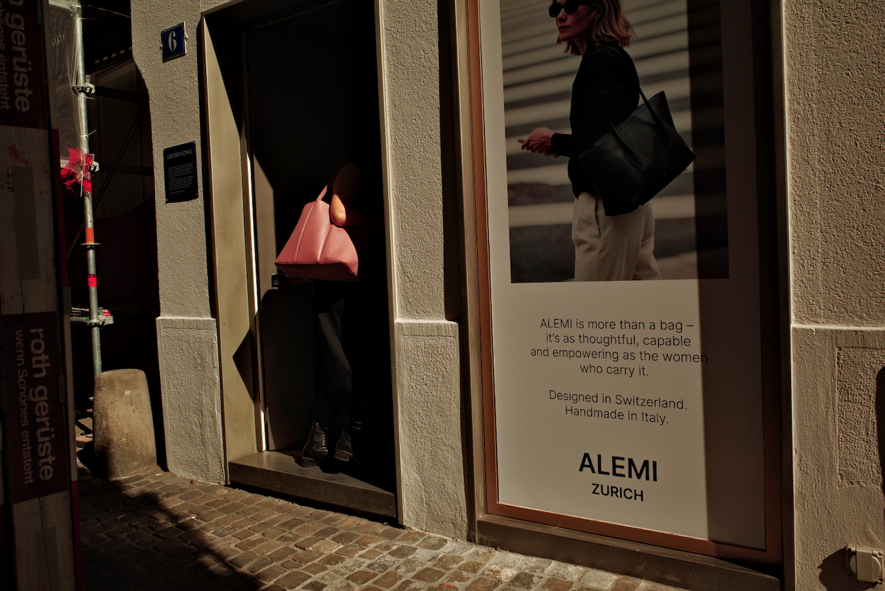 A person carrying a pink bag is entering a building next to a large advertisement for Alemi Zurich, describing the brand’s bags as thoughtful and empowering. (Zurich, 2025)