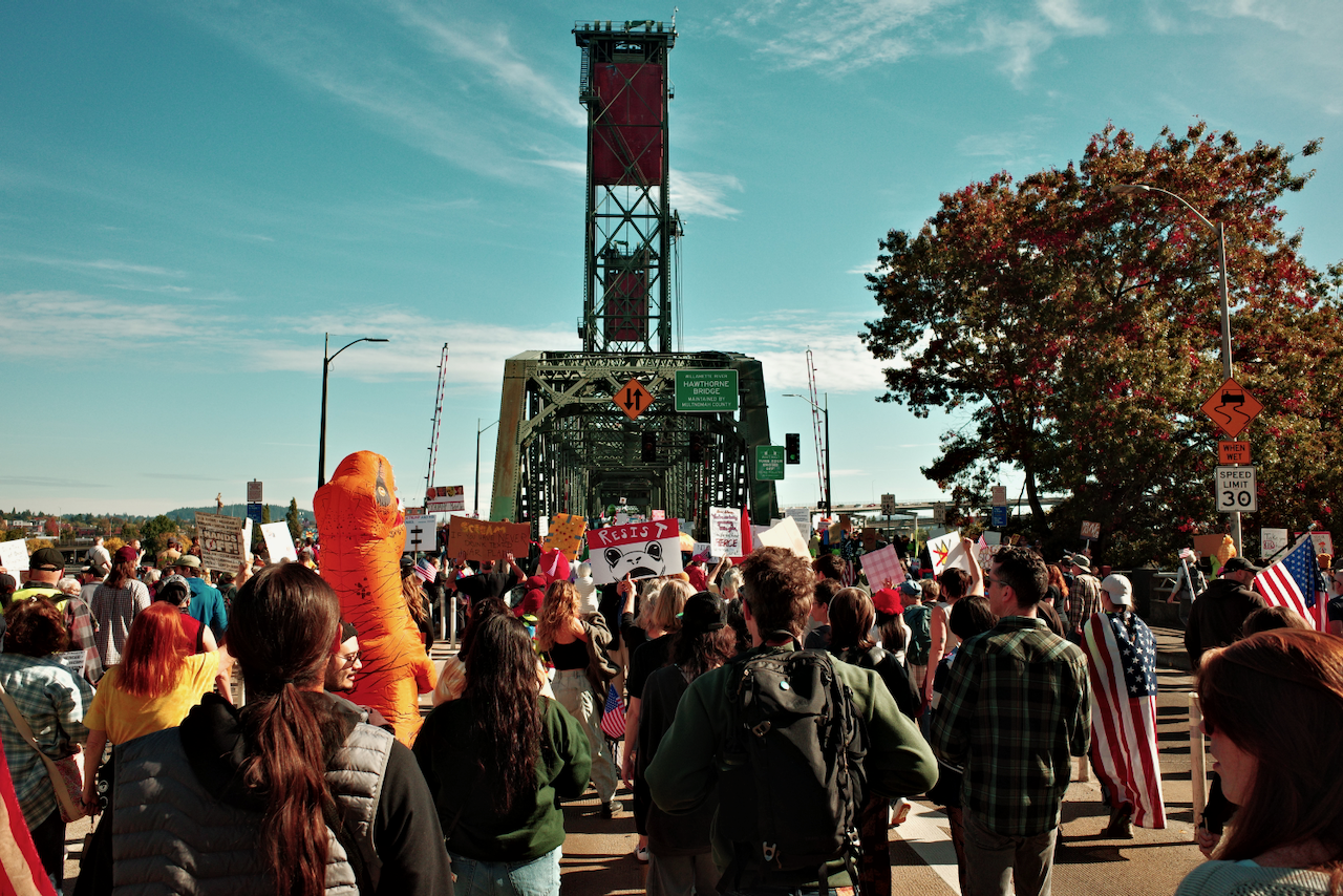 A crowd of people holding signs is marching on a street toward a bridge under a clear blue sky. (Portland, 2025)