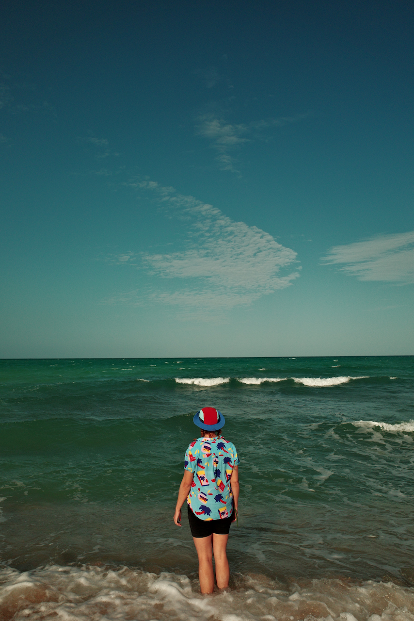 A person is standing at the edge of the ocean, wearing a colorful shirt and hat, looking toward the horizon. (Mexico, 2025)