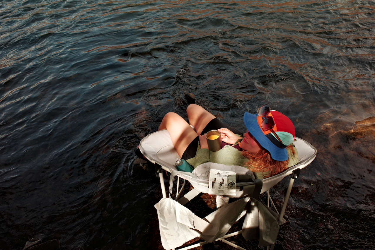 A person relaxes in a reclining chair near the water, wearing a colorful hat and holding a mug. (Maupin, 2026)