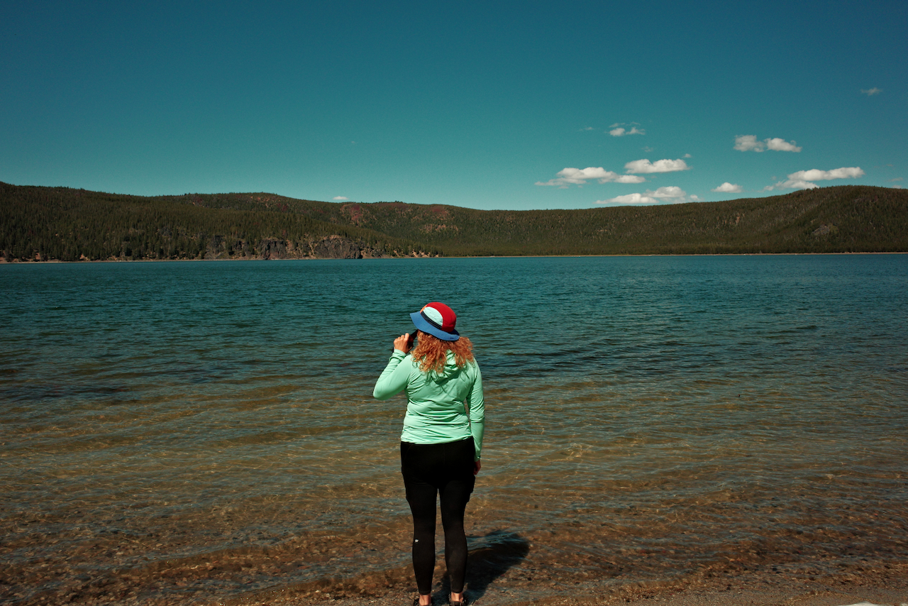 A person wearing a mint green sun shirt and a red, white, and blue hat stands at the edge of a calm lake with forested hills in the background under a clear blue sky.