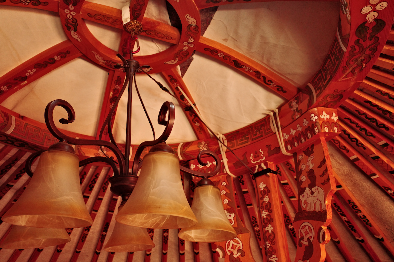 A photo from inside of the yurt, showing the roof’s central ring and a set of light’s hanging down from it. The wood structural pieces are decorated with intricate, hand drawn paintings in a traditional Mongolian style.