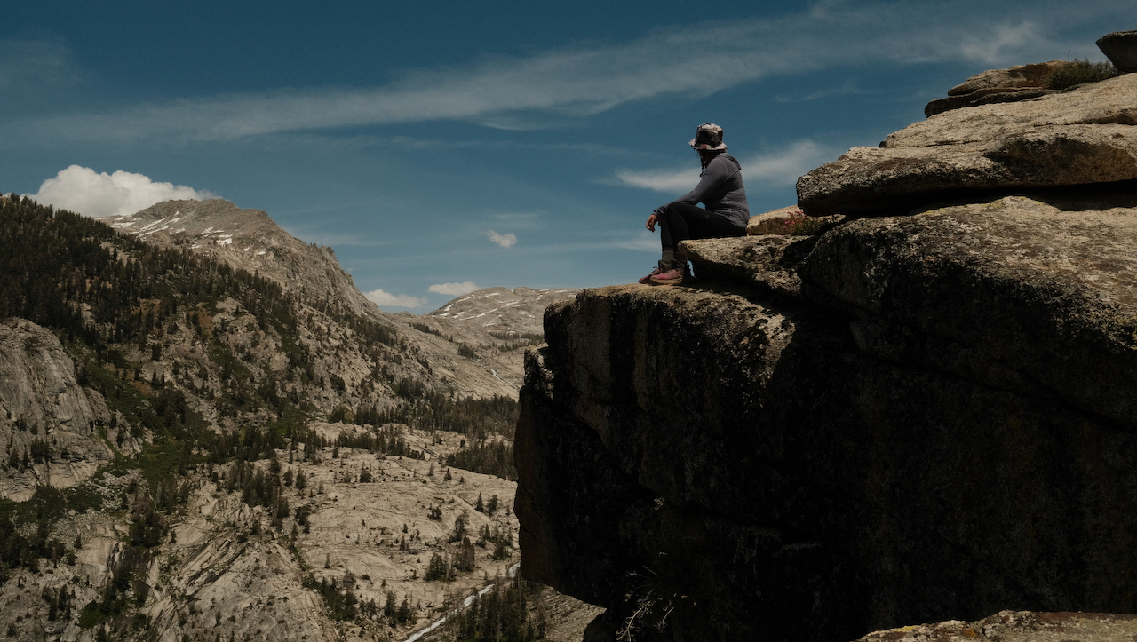 A photo of a woman sitting on a road ledge, overlooking a valley.