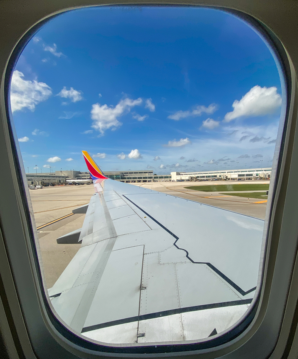 A view from an airplane window shows the wing of the aircraft with a clear blue sky and scattered clouds visible above the airport tarmac.