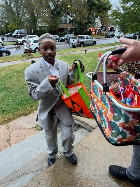A child dressed in a gray suit is holding a Halloween bag while receiving candy from an adult's basket on the front steps of a house.