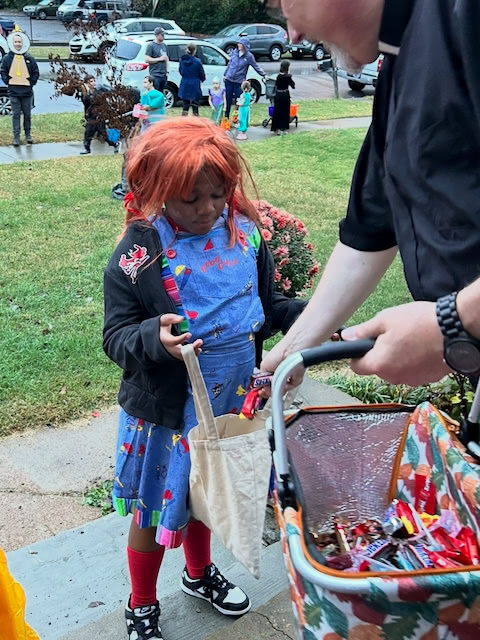 A child dressed in a colorful Halloween costume is receiving candy from an adult outside.