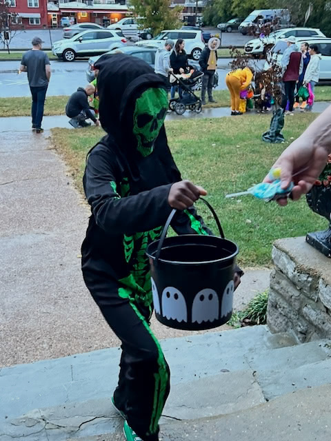 A child dressed in a green and black skeleton costume is trick-or-treating, holding a bucket with ghost designs as they receive candy.