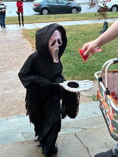 A child dressed in a black hooded costume with a white ghostly mask receives candy on Halloween.
