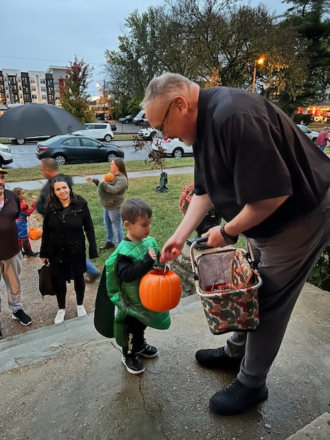 A young child in a dinosaur costume is receiving candy from an adult during what appears to be trick-or-treating.