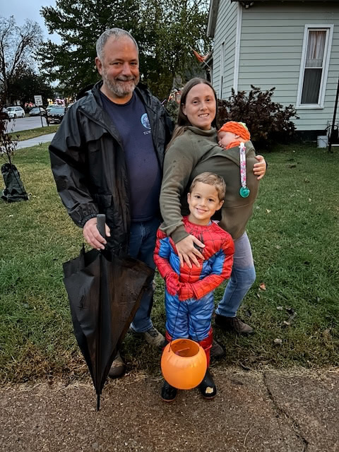 A family is gathered outside, with a child dressed as Spider-Man and a baby wearing a pumpkin hat, ready for Halloween.