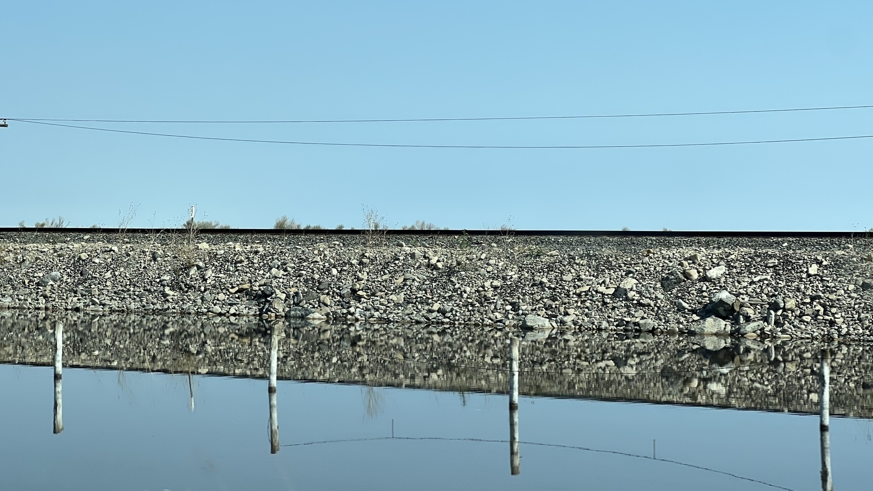 Side view of raised train tracks reflected in still water in front of the embankment.