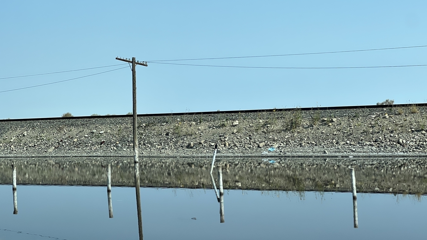 Side view of raised train tracks reflected in still water in front of the embankment.