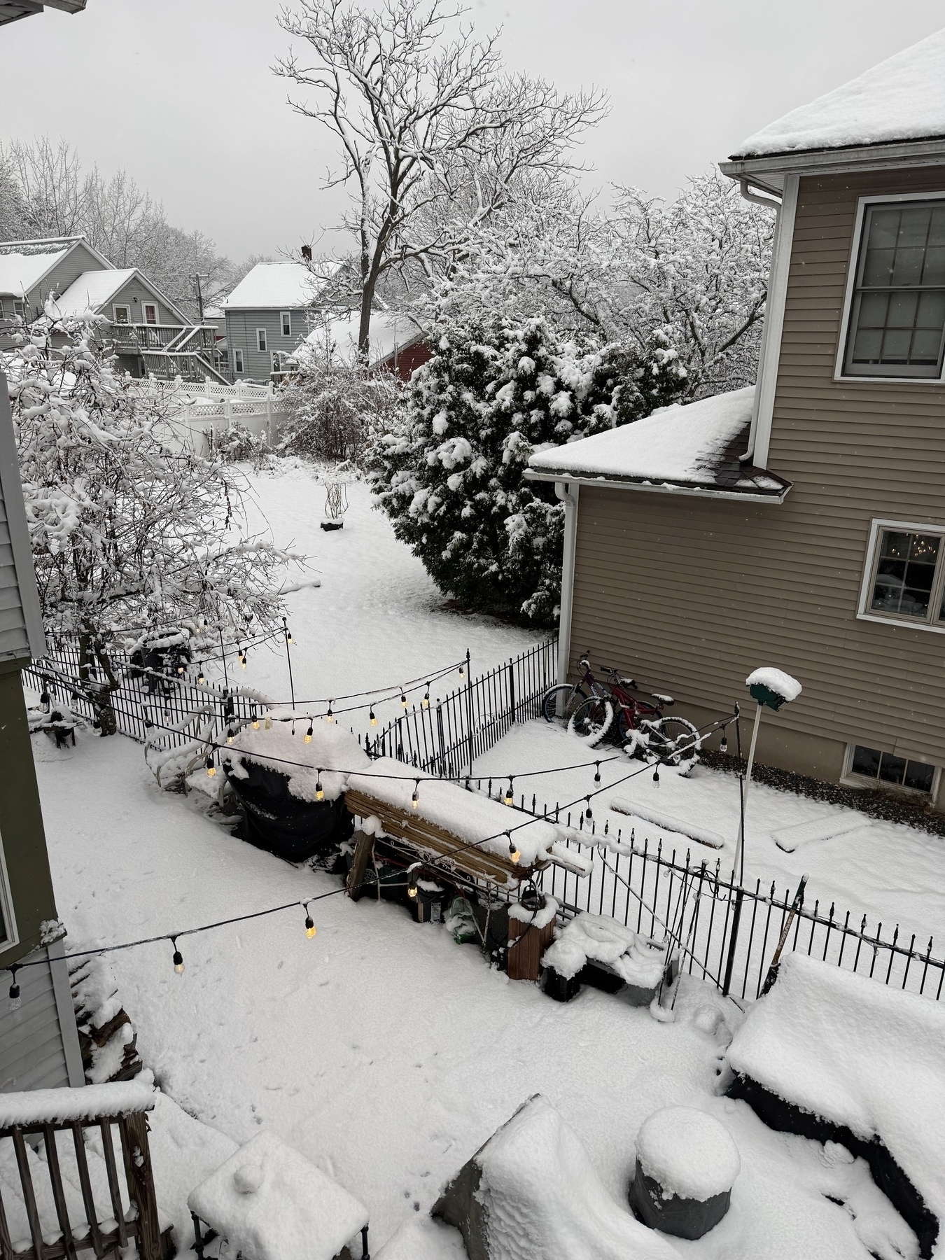 A snow-covered backyard features a patio with string lights, bicycles, and surrounding trees and houses blanketed in snow.