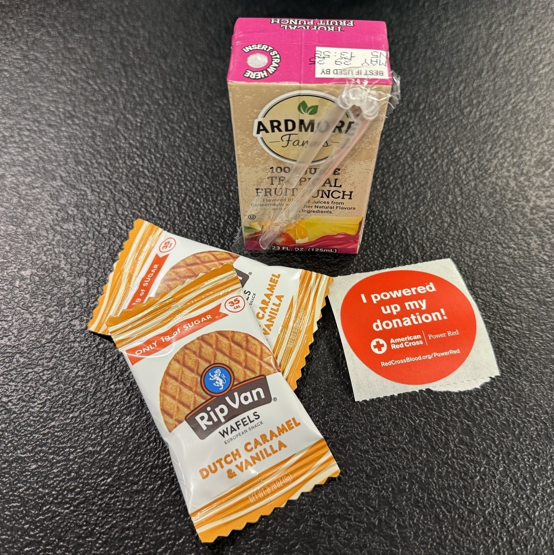 A carton of Ardmore Farm Tropical Fruit Punch, a sticker related to blood donation, and two Rip Van Wafels Dutch Caramel & Vanilla snacks are placed on a dark surface.